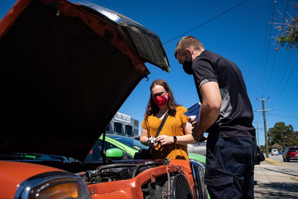 A friendly voltaic employee inspecting the engine of a car with the happy customer by his side.